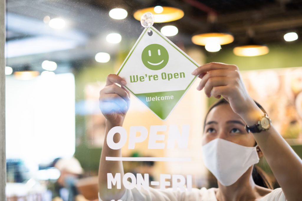 Business owner wear protective face mask ppe hanging open sign at her restaurant / café, open again after lock down due to outbreak of coronavirus covid-19